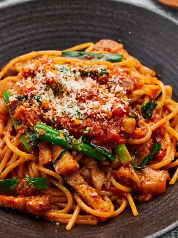 Spicy and savory 15 minute Pork & Kimchi Pasta piled high on a dark plate, the vibrant red of the kimchi and juicy pork belly pops against the creamy strands of spaghetti and vibrant green scallions.
