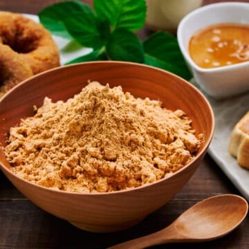 Close-up shot of a wooden bowl filled with homemade kinako powder, its golden hue hinting at its rich, nutty flavor. With kinako doughnuts and kinako butter in the background.