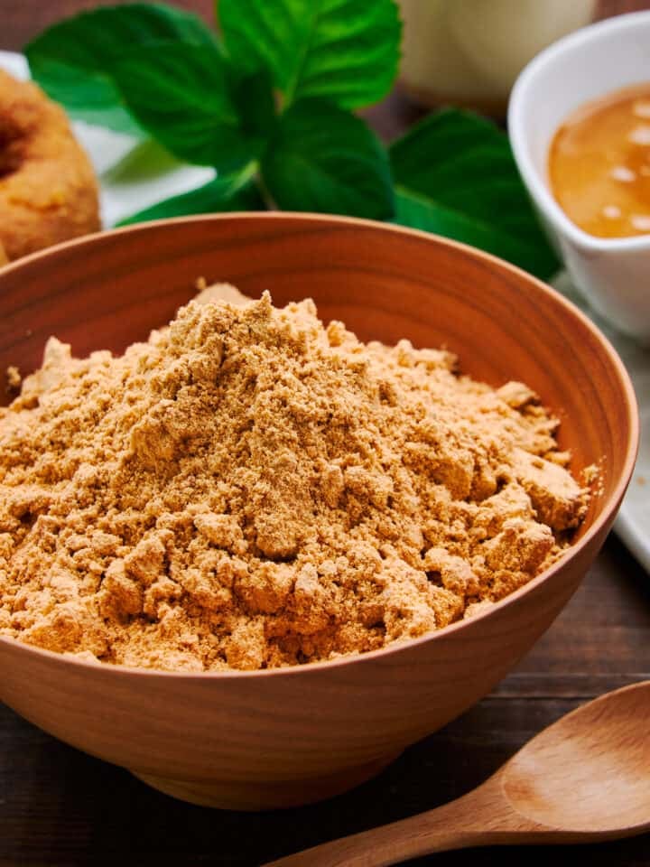 Close-up shot of a wooden bowl filled with homemade kinako powder, its golden hue hinting at its rich, nutty flavor. With kinako doughnuts and kinako butter in the background.