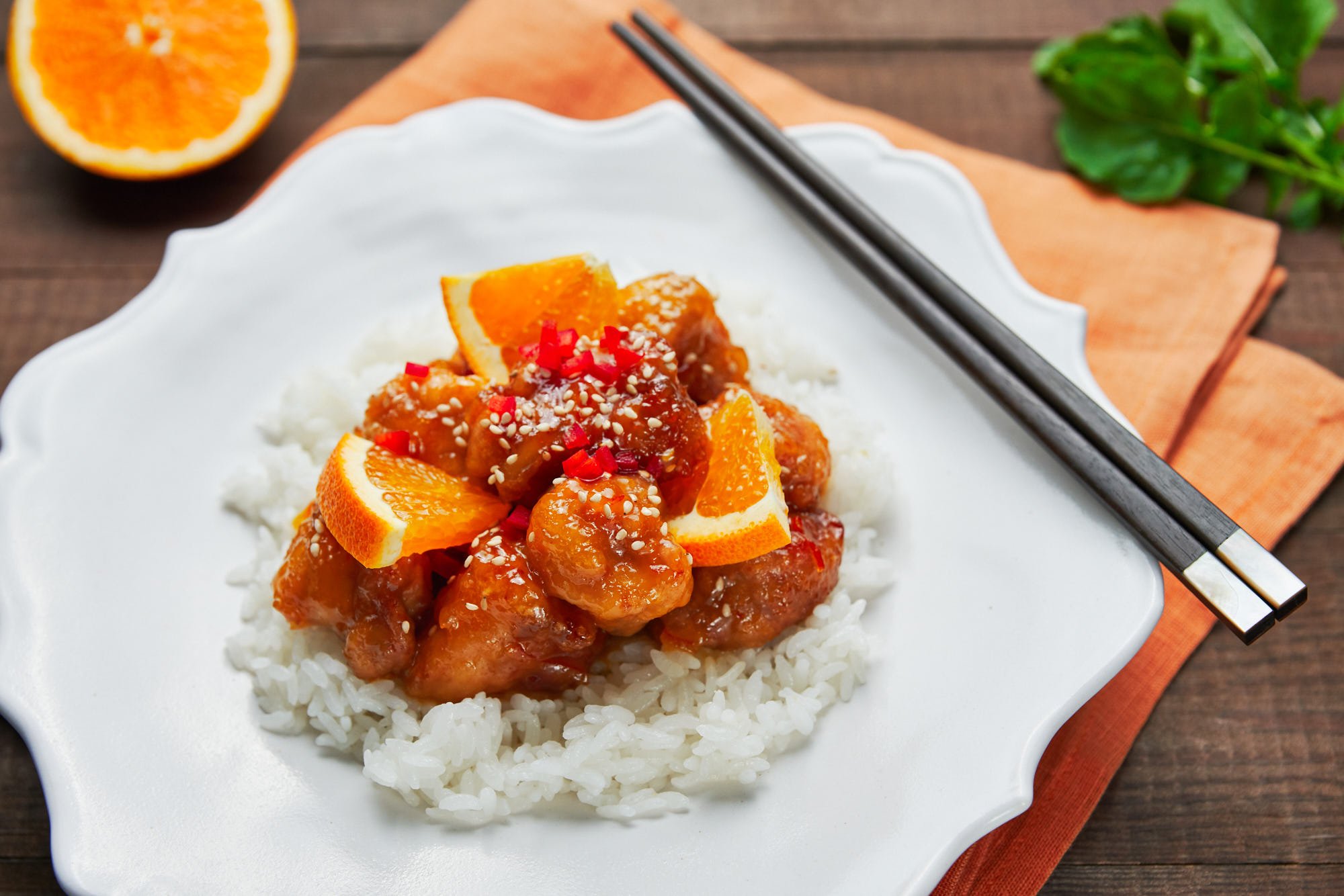 Orange chicken with orange segments served over a bed of white rice on a white plate with chopsticls.