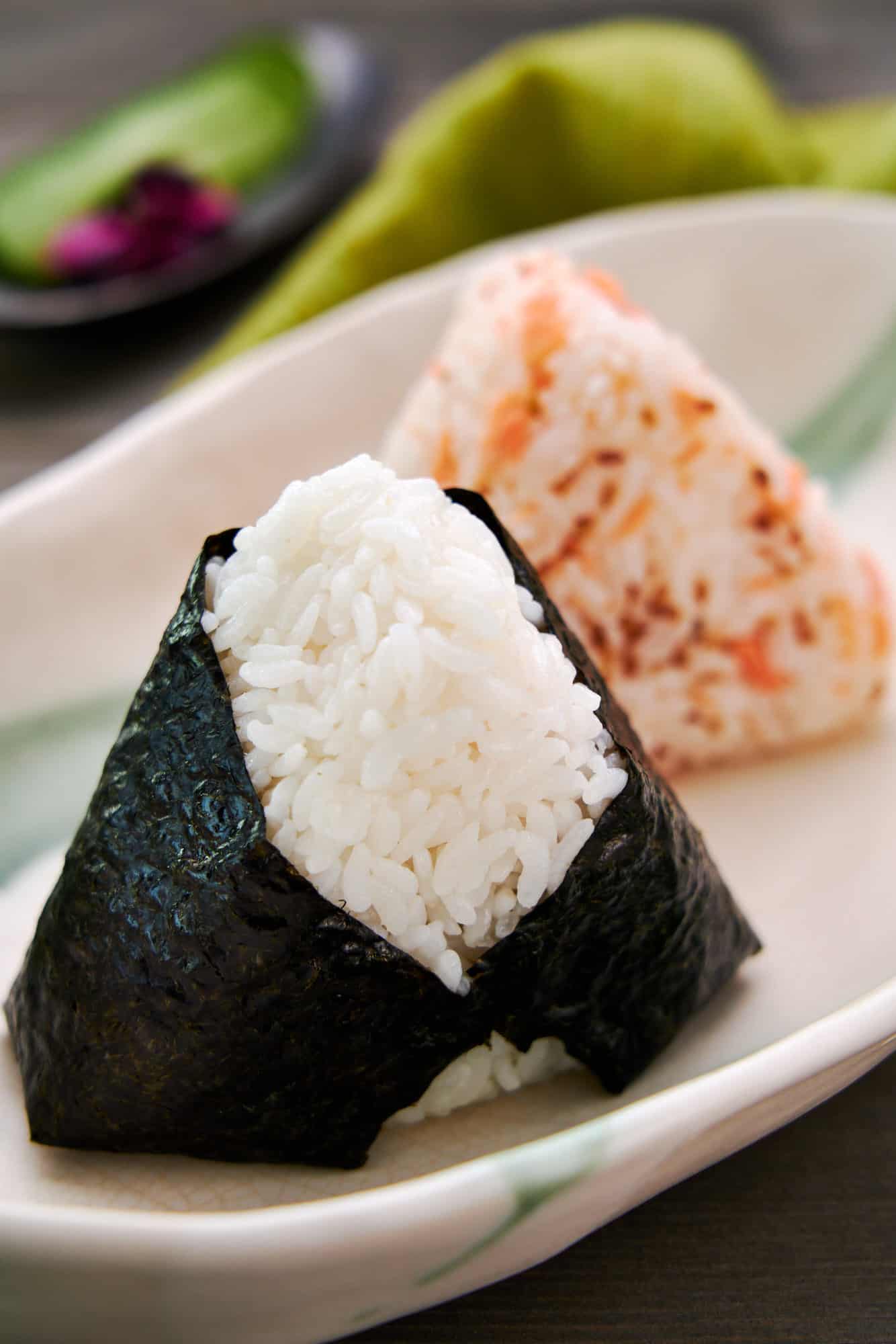 Two ways to make salmon onigiri. A traditional salted salmon flake onigiri wrapped in nori in the foreground and a rice ball mixed with salmon flakes and toasted sesame in the background.