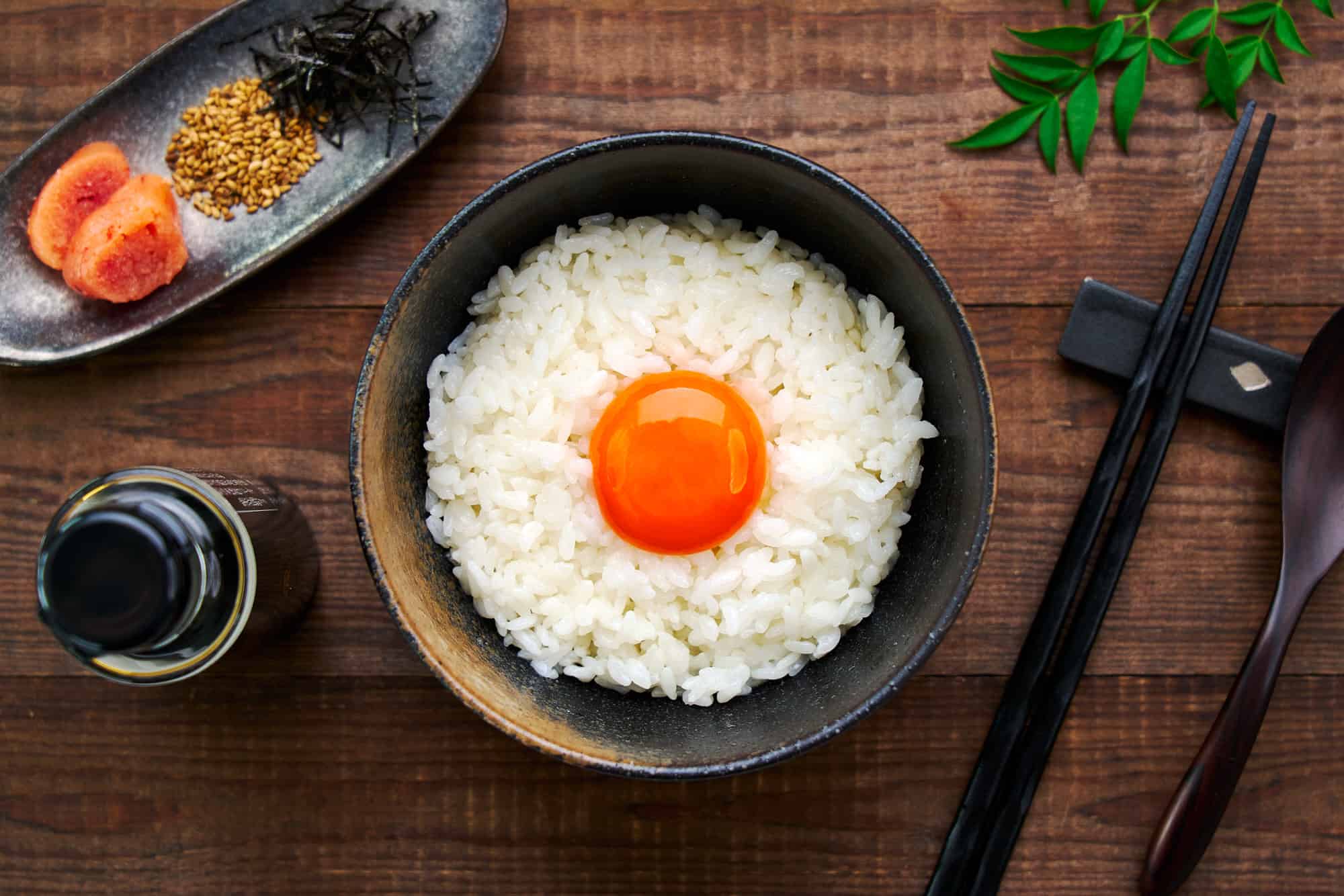 Overhead shot of a bowl of creamy Tamago Kake Gohan, showing the vibrant egg yolk resting atop a bed of fluffy white rice. With soy sauce and various mix-ins on the side.